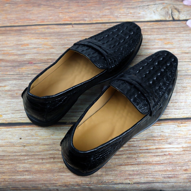 May include: A pair of black leather loafers with a crocodile skin texture. The shoes have a slip-on design with a low heel and a tan interior. The loafers are displayed on a wooden surface.