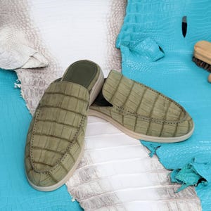 May include: A pair of olive green crocodile-textured leather slip-on shoes with a light beige sole. The shoes are displayed on a backdrop of white and turquoise crocodile-textured leather.