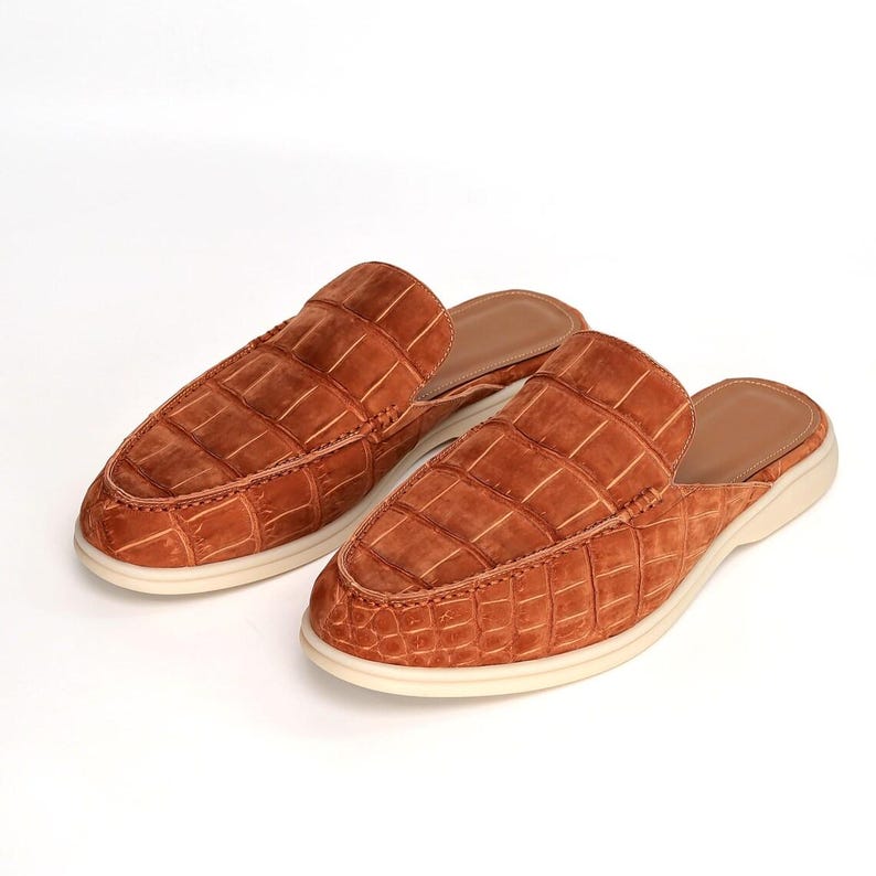 May include: A pair of brown leather slip-on shoes with a crocodile pattern. The shoes have a rounded toe and a cream-colored sole. The open-back design offers a casual yet stylish look.