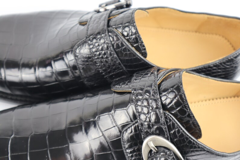 May include: Two black leather shoes with a crocodile skin pattern and silver buckles. The shoes have a pointed toe and a low heel.