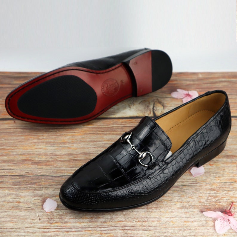 May include: Black leather loafers with a crocodile pattern and a silver buckle detail. The loafers have a pointed toe and a low heel.