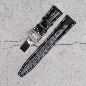 May include: Black leather watch strap with a silver buckle. The strap has a crocodile pattern and is designed for a watch with a deployment clasp.