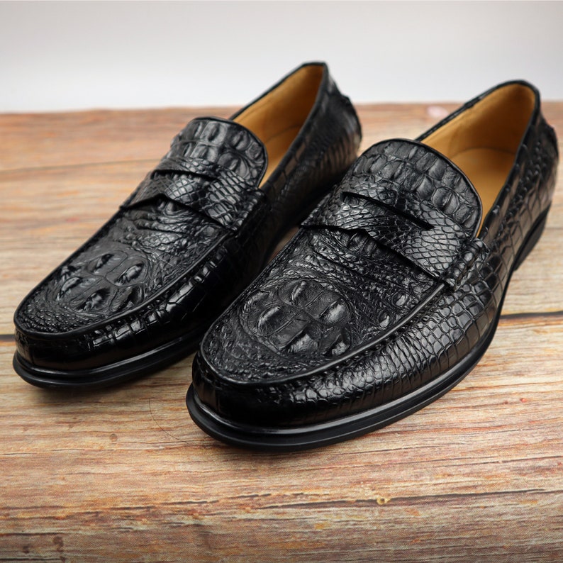 May include: A pair of black leather loafers with a crocodile skin texture. The shoes have a classic design with a rounded toe and a strap across the top. The soles are black and the shoes are displayed on a wooden surface.