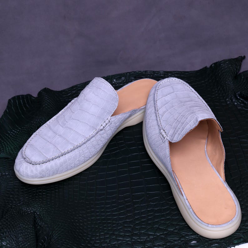 May include: A pair of light blue leather slip-on shoes with a crocodile skin texture. The shoes have a beige interior and a white sole. The shoes are displayed on a dark green textured surface.