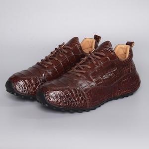 Puede incluir: Un par de zapatillas de cuero marr&oacute;n con textura de piel de cocodrilo. Los zapatos tienen cordones marrones, un cuello acolchado y una suela negra. Las zapatillas se muestran sobre un fondo gris claro.