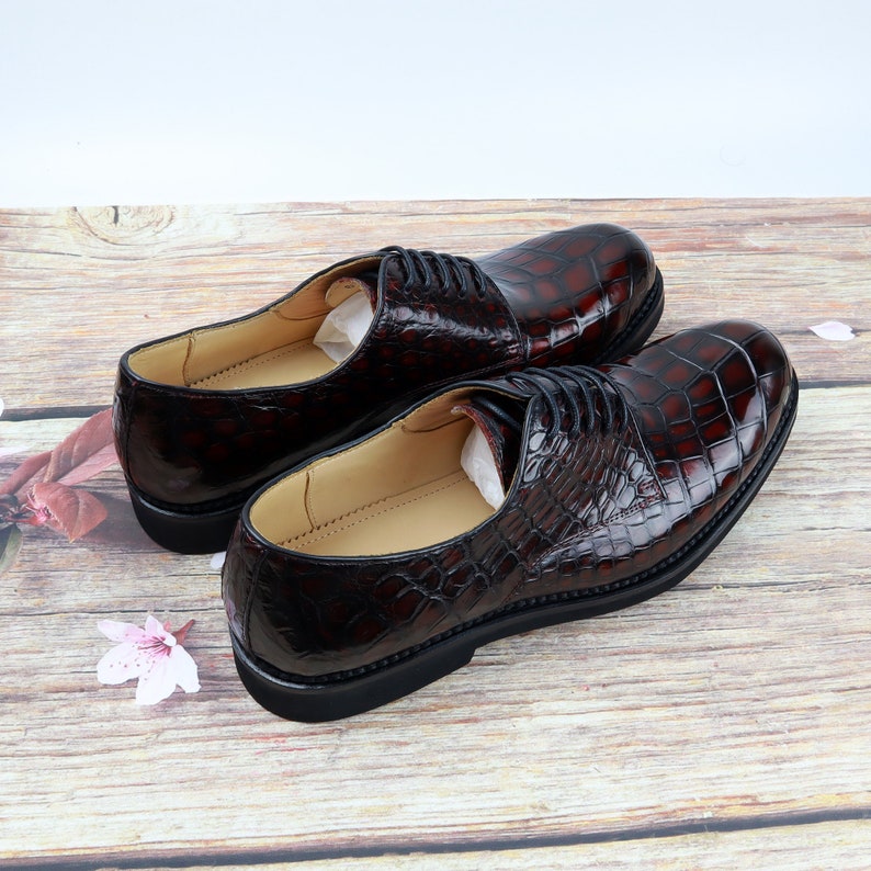 Peut inclure: Une paire de chaussures habill&eacute;es en cuir marron avec un motif crocodile. Les chaussures ont des lacets et sont sur une surface en bois.