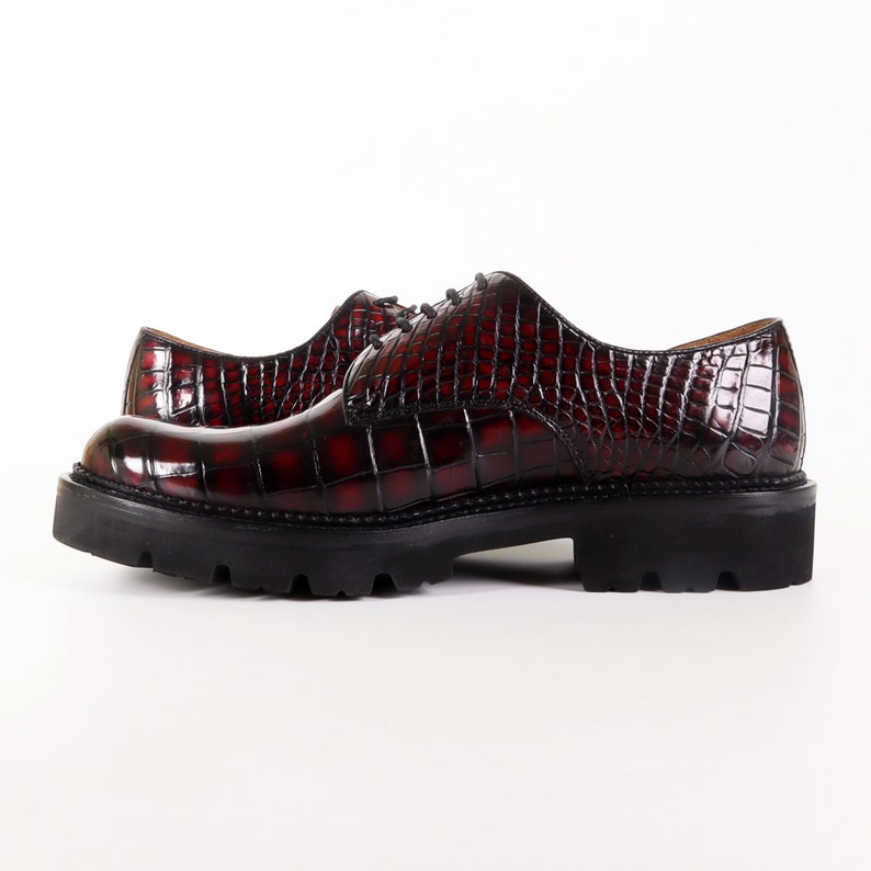 May include: A pair of burgundy leather lace-up shoes with a crocodile-embossed pattern. The shoes have a chunky black rubber sole.