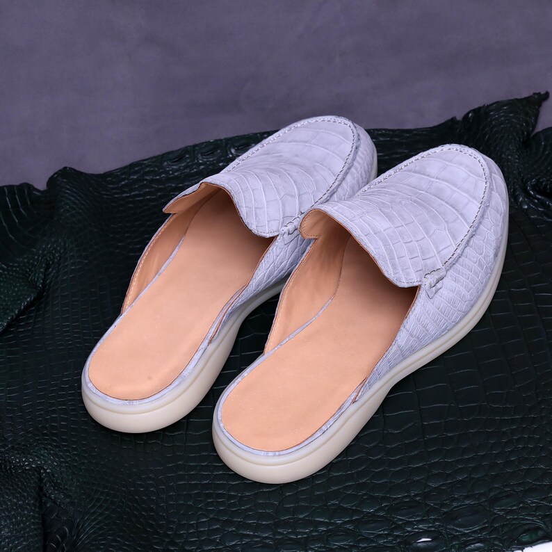 May include: A pair of light blue crocodile-textured leather loafers. The slip-on shoes have a rounded toe, a beige interior, and a cream-colored sole. The shoes are displayed on a dark green textured surface.