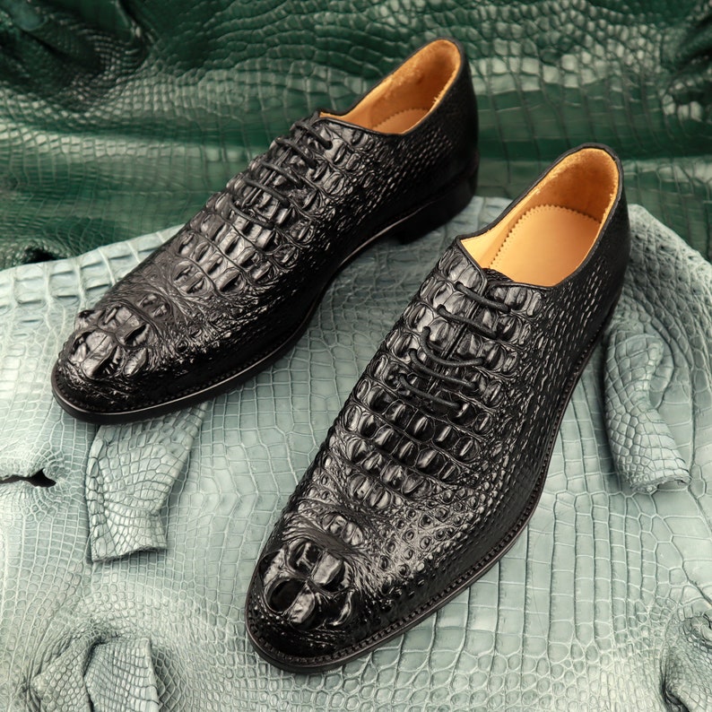 May include: A pair of black leather dress shoes with crocodile embossed detailing. The shoes have lace-up closures and are sitting on a green and blue textured surface.