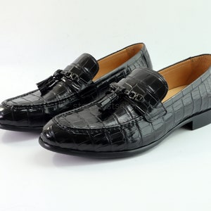May include: A pair of black leather loafers with a crocodile pattern and tassel accents. The loafers feature a metal chain detail on the front.