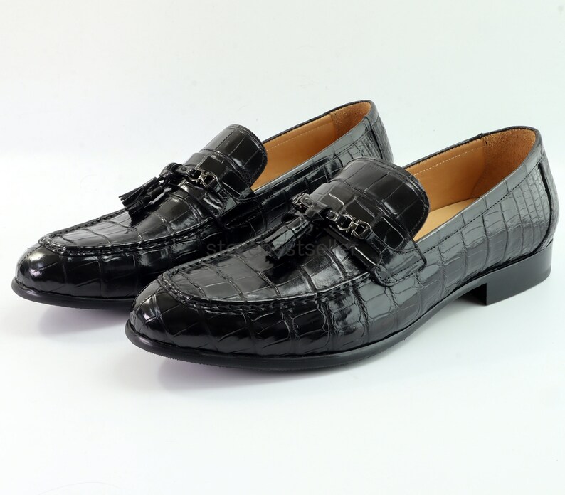 May include: A pair of black leather loafers with a crocodile pattern and tassel accents. The loafers feature a metal chain detail on the front.