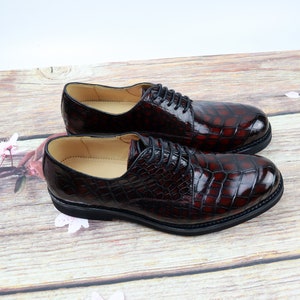 Peut inclure: Une paire de chaussures habill&eacute;es en cuir bordeaux &agrave; motif crocodile. Les chaussures ont des lacets noirs et une semelle en caoutchouc noire.