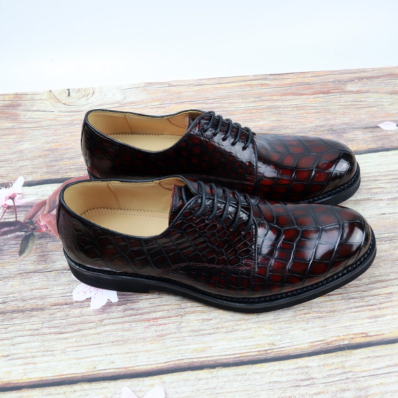 Peut inclure: Une paire de chaussures habill&eacute;es en cuir bordeaux &agrave; motif crocodile. Les chaussures ont des lacets noirs et une semelle en caoutchouc noire.