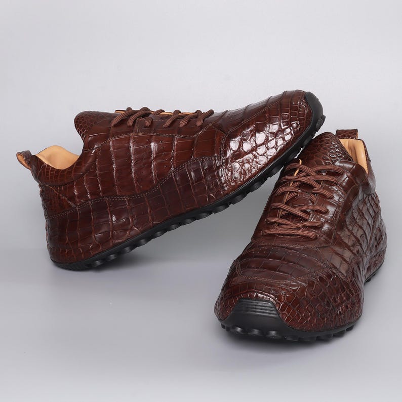 Puede incluir: Un par de zapatillas de cuero marr&oacute;n con textura de piel de cocodrilo. Los zapatos tienen cordones marrones y suelas negras. El interior del zapato es de color beige claro.