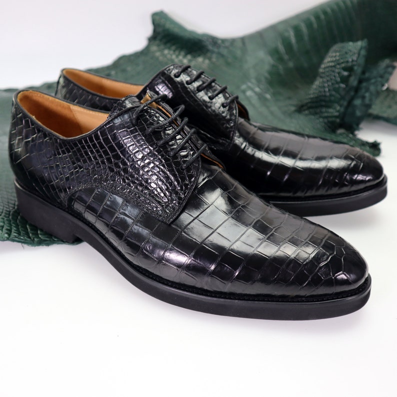 K&ouml;nnte beinhalten: Ein Paar schwarze Lederschuhe mit Kroko-Muster. Die Schuhe haben einen Schn&uuml;rverschluss und eine spitze Zehenpartie.