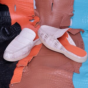 May include: A pair of white crocodile-textured leather slip-on shoes with a light-colored sole. The interior is orange. The shoes are displayed on a backdrop of various colored crocodile leather hides, including black, orange, brown, and blue.