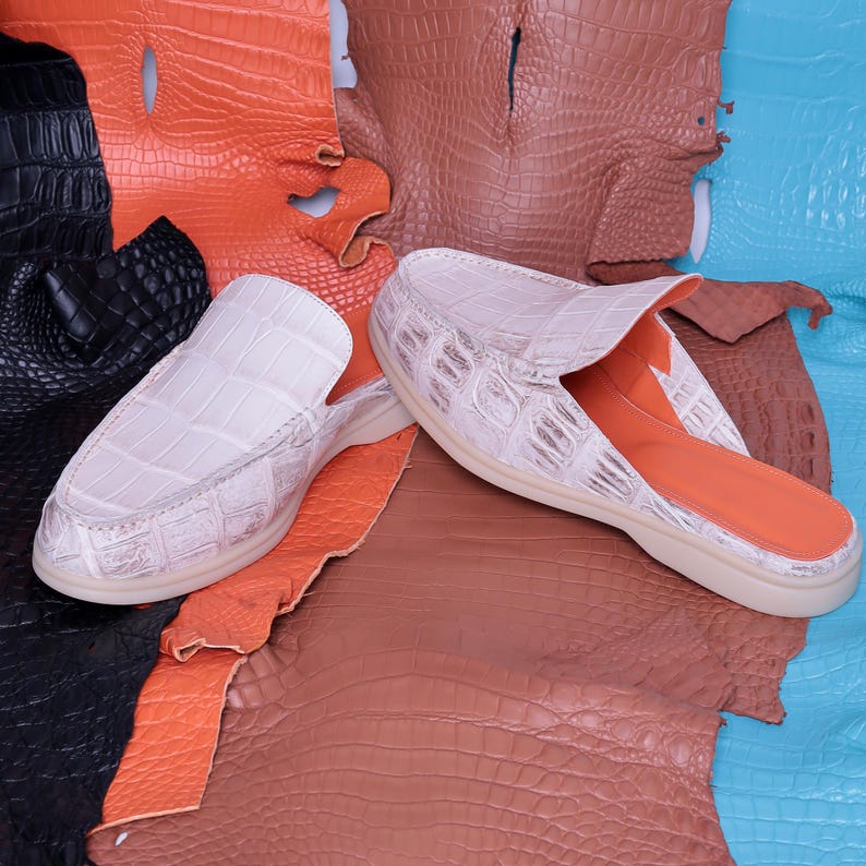May include: A pair of white crocodile-textured leather slip-on shoes with a light-colored sole. The interior is orange. The shoes are displayed on a backdrop of various colored crocodile leather hides, including black, orange, brown, and blue.