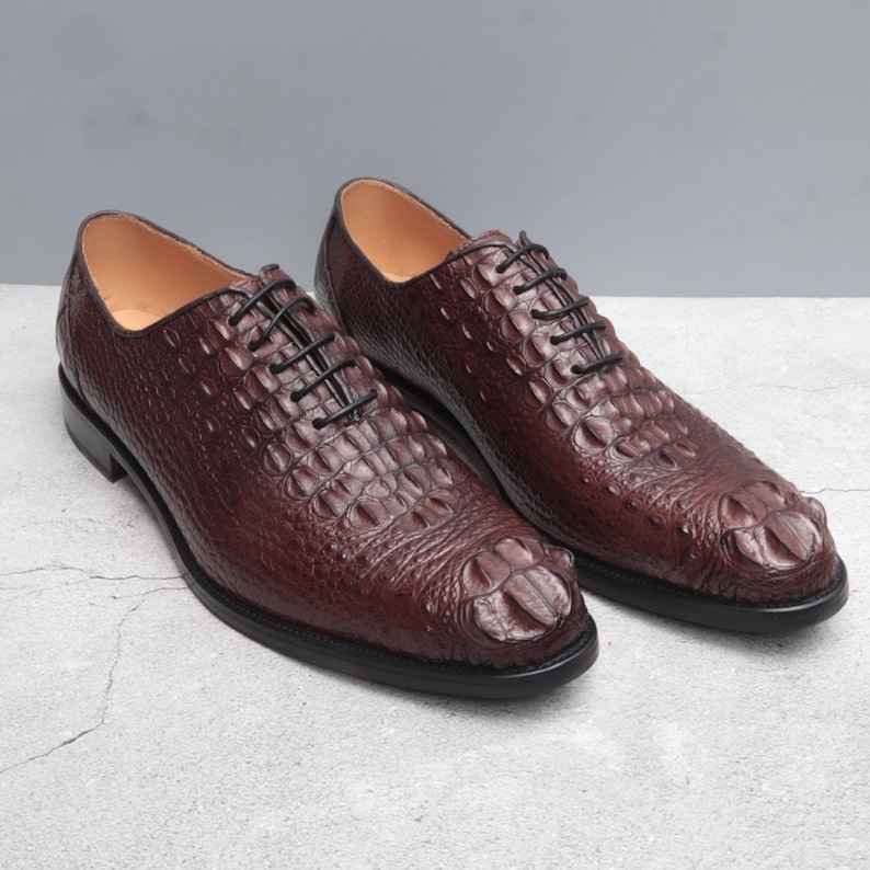 Pair of dark brown men’s wholecut shoes displayed on light concrete surface, Alligator shoes men , black waxed laces, closed lacing, and rounded square toe with leather sole.