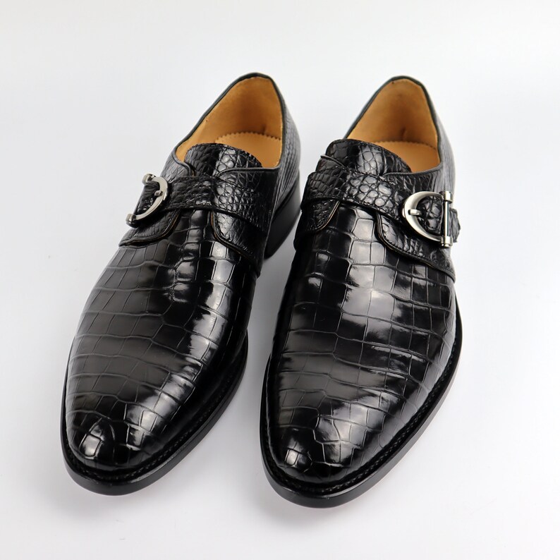 May include: A pair of black leather dress shoes with a crocodile pattern. The shoes have a buckle fastening and a pointed toe.