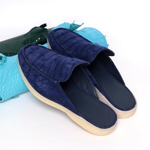 May include: Navy blue slip-on shoes with a crocodile-effect texture. The shoes have a suede-like finish and a cream-coloured sole. The background includes pieces of green and blue leather.