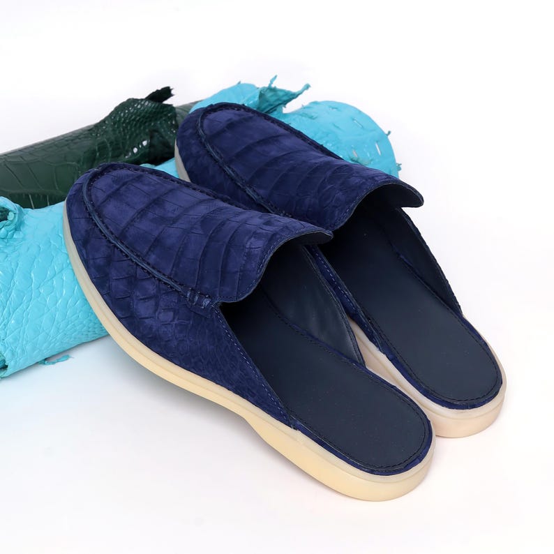 May include: Navy blue slip-on shoes with a crocodile-effect texture. The shoes have a suede-like finish and a cream-coloured sole. The background includes pieces of green and blue leather.