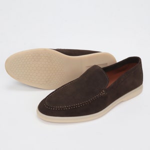 Op de afbeelding: Een paar bruine su&egrave;de loafers met een witte rubberen zool. De loafers hebben een slip-on ontwerp en een gestikt detail rond de teen.