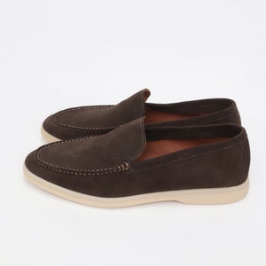 Op de afbeelding: Een paar bruine su&egrave;de loafers met een witte rubberen zool. De loafers hebben een slip-on ontwerp en een gestikt detail rond de neus.