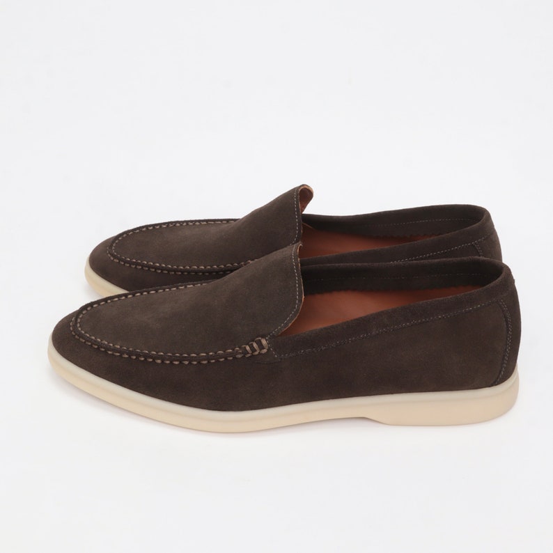 Op de afbeelding: Een paar bruine su&egrave;de loafers met een witte rubberen zool. De loafers hebben een slip-on ontwerp en een gestikt detail rond de neus.