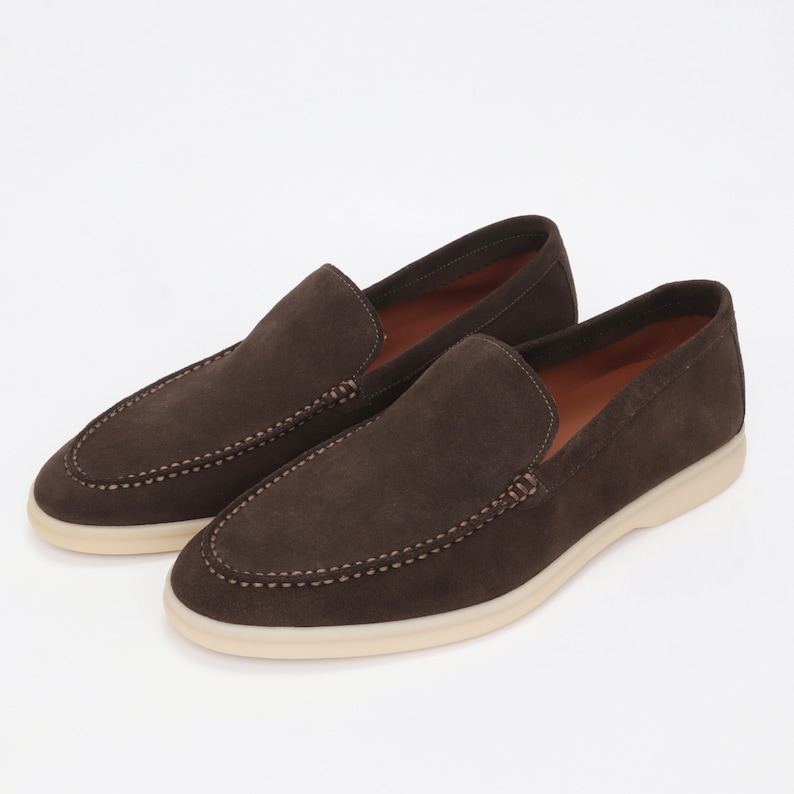 Op de afbeelding: Een paar bruine su&egrave;de loafers met een witte rubberen zool. De loafers hebben een gestikt detail rond de rand van de zool.