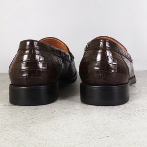 Puede incluir: Un par de mocasines de cuero marr&oacute;n con un patr&oacute;n de cocodrilo en relieve. Los mocasines tienen una punta redonda y un tac&oacute;n bajo.