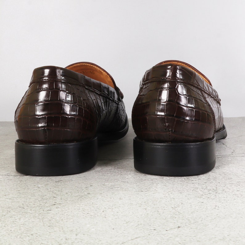 Puede incluir: Un par de mocasines de cuero marr&oacute;n con un patr&oacute;n de cocodrilo en relieve. Los mocasines tienen una punta redonda y un tac&oacute;n bajo.