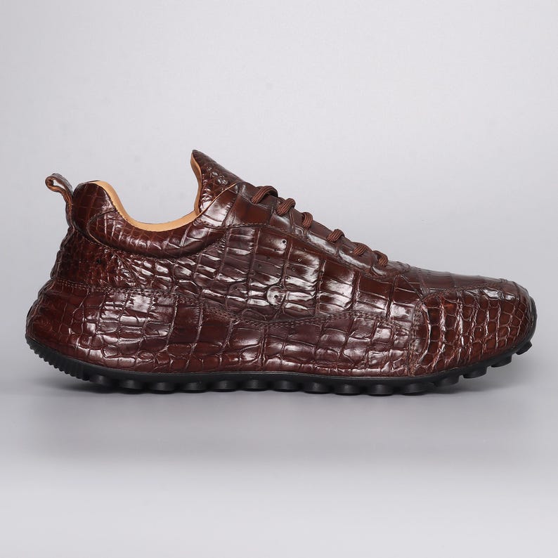 Puede incluir: Una zapatilla de cuero marr&oacute;n con textura de piel de cocodrilo. El zapato tiene una puntera redondeada, cordones marrones y una suela negra. La zapatilla est&aacute; dise&ntilde;ada para uso casual.