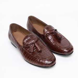 May include: A pair of brown leather loafers with a crocodile skin texture. Each shoe features a decorative tassel and a rounded toe. The shoes are set against a white background.