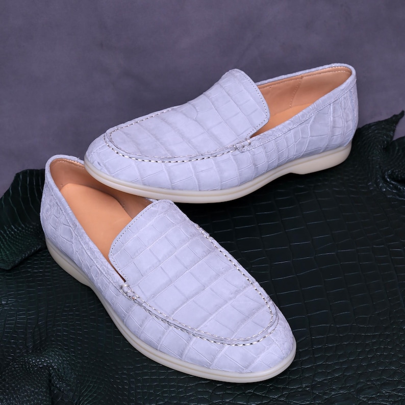 May include: A pair of light blue leather loafers with a crocodile skin texture. The shoes have a rounded toe, a stitched vamp, and a cream-colored sole. The interior is a light tan color.