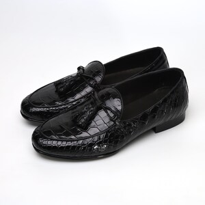 May include: A pair of glossy black leather loafers with a crocodile texture. Each shoe features a decorative tassel and a rounded toe. The shoes are displayed on a white background.