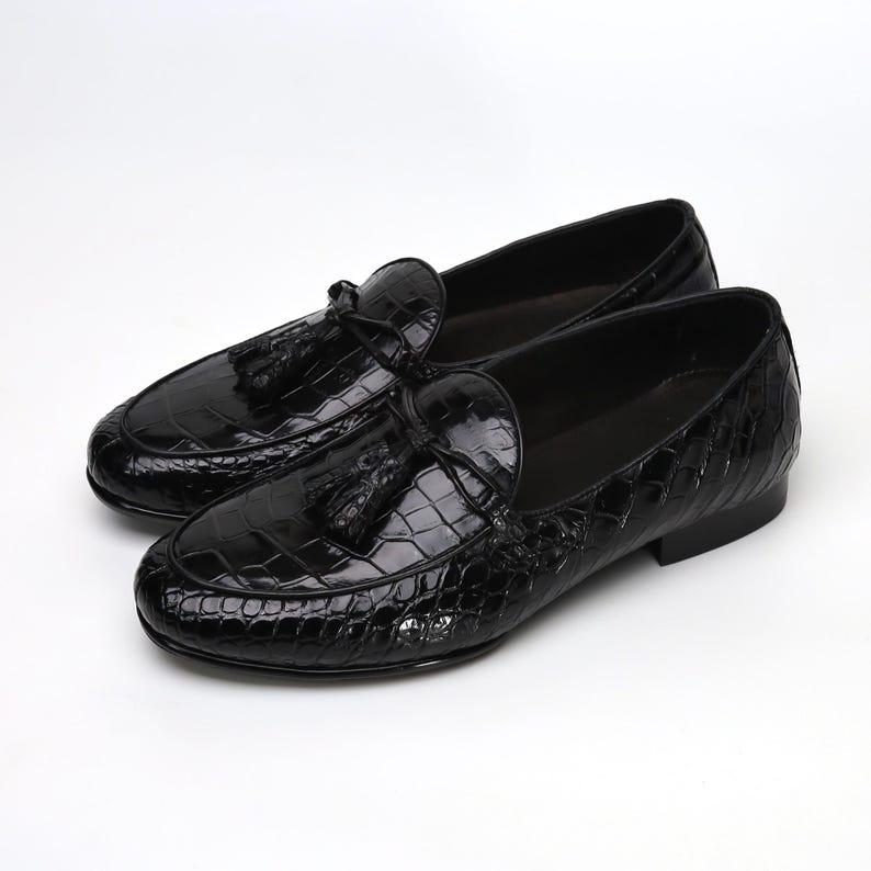May include: A pair of glossy black leather loafers with a crocodile texture. Each shoe features a decorative tassel and a rounded toe. The shoes are displayed on a white background.