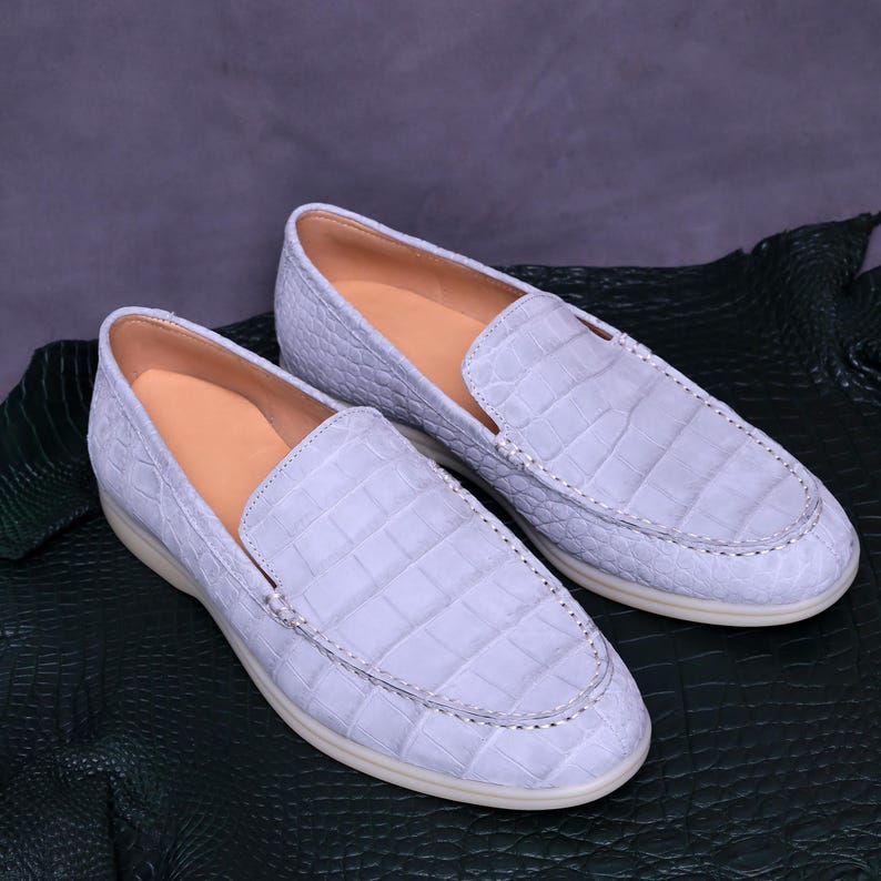 May include: A pair of light blue leather loafers with a crocodile skin texture. The shoes have a rounded toe, a light-colored sole, and a tan interior. The shoes are displayed on a dark green surface.