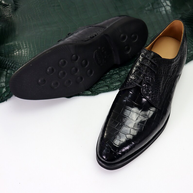 K&ouml;nnte beinhalten: Ein Paar schwarze Lederschuhe mit Kroko-Muster. Die Schuhe haben einen Schn&uuml;rverschluss und eine spitze Zehenpartie.