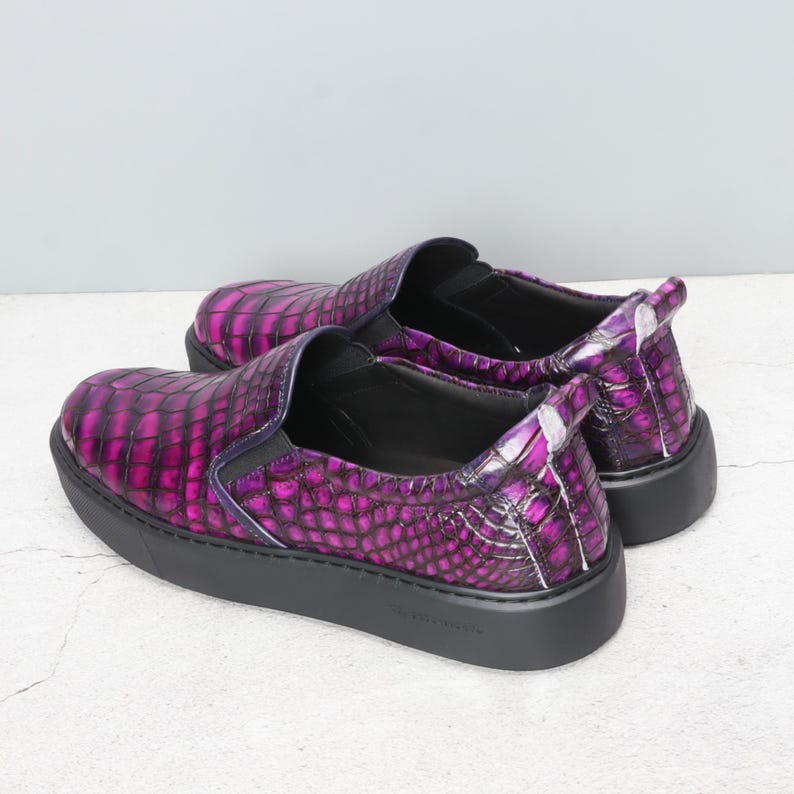 May include: A pair of slip-on shoes with a textured, purple and black pattern. The shoes have a black sole and a rounded toe. The design resembles crocodile skin. The shoes are set against a neutral background.