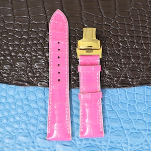 Peut inclure: Deux bracelets de montre en alligator rose avec une boucle dorée. Les bracelets sont sur un fond brun et bleu.