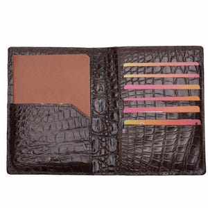 Può includere: Portafoglio in pelle marrone aperto con una texture a pelle di coccodrillo. Il portafoglio ha più fessure per carte con carte colorate e una tasca per un passaporto o un taccuino. Il portafoglio è aperto, mostrando l'interno.