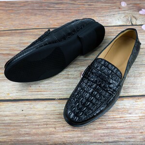 May include: Pair of black leather loafers with a crocodile skin texture. One shoe is right-side up, and the other is upside down, showing the sole. The shoes are on a wooden surface.