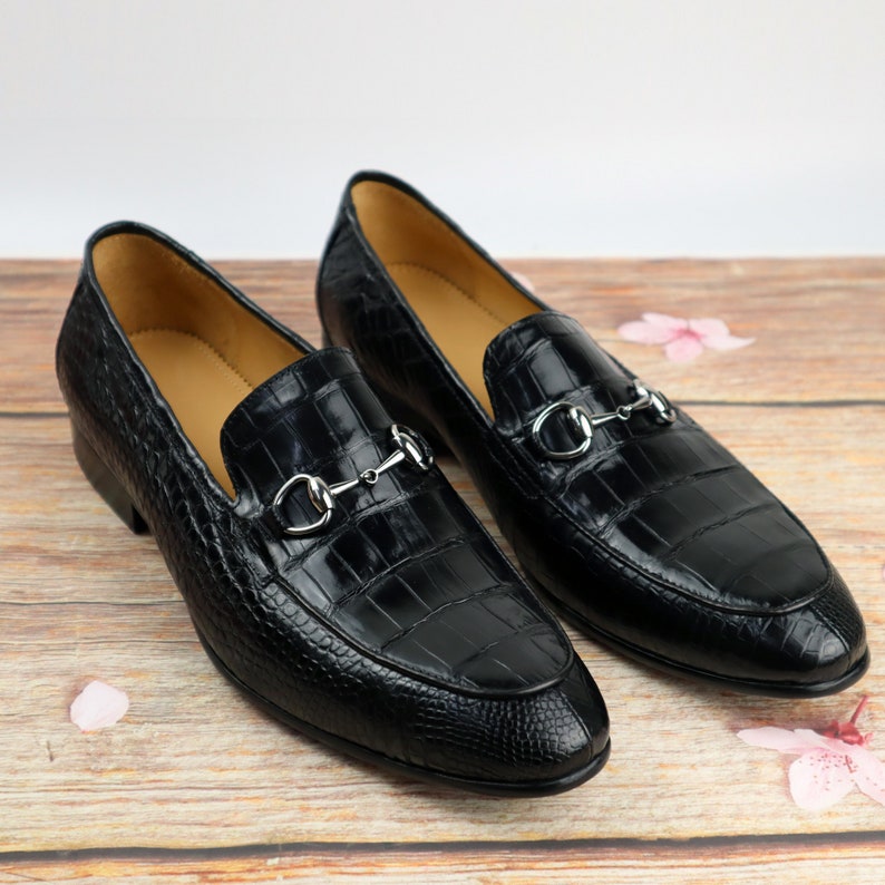 May include: A pair of black leather loafers with a crocodile pattern and silver metal buckles. The loafers are on a wooden surface.