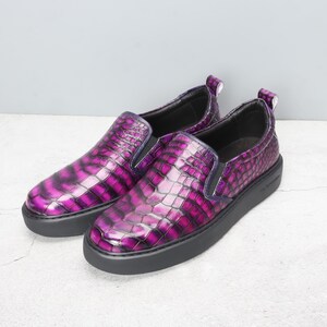 May include: A pair of slip-on shoes with a glossy, textured, purple and black crocodile pattern. The shoes have a black sole and a small pull tab at the heel. The design is eye-catching and modern.