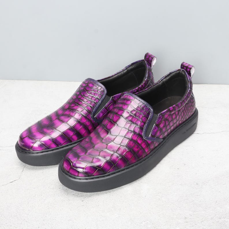 May include: A pair of slip-on shoes with a glossy, textured, purple and black crocodile pattern. The shoes have a black sole and a small pull tab at the heel. The design is eye-catching and modern.