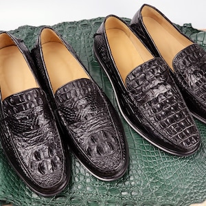 May include: Four pairs of black leather loafers with a crocodile skin texture are displayed on a green textured surface. The shoes have a classic design with a rounded toe and a low heel. The leather has a glossy finish.