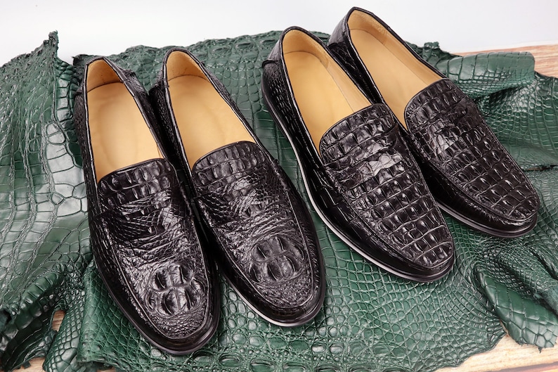 May include: Four pairs of black leather loafers with a crocodile skin texture are displayed on a green textured surface. The shoes have a classic design with a rounded toe and a low heel. The leather has a glossy finish.