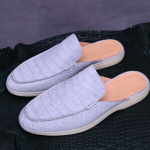 May include: A pair of light blue slip-on shoes with a crocodile skin texture. The shoes have a rounded toe, a tan insole, and a cream-colored sole. The shoes are displayed on a dark green textured surface.