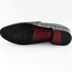 May include: The underside of a black leather loafer with a crocodile pattern. The sole is black rubber with a textured pattern. The shoe has a red leather heel and a brown leather insole with the text "Goodyear Welt" and a winged horse logo.