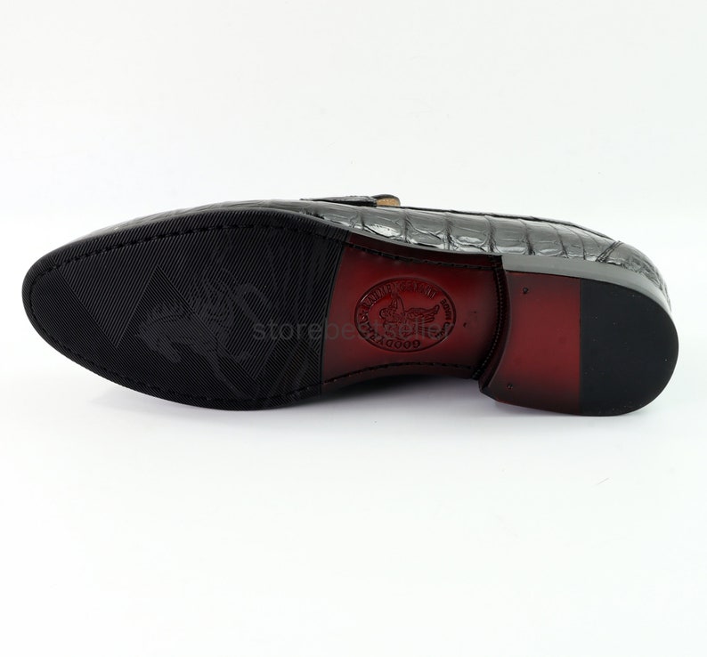 May include: The underside of a black leather loafer with a crocodile pattern. The sole is black rubber with a textured pattern. The shoe has a red leather heel and a brown leather insole with the text "Goodyear Welt" and a winged horse logo.